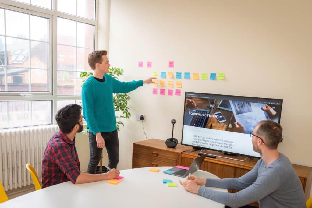 Two male employees sat at a desk in a meeting room looking at a screen and another male employee stood up pointing to a post it note