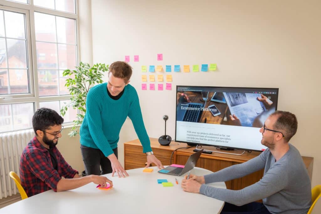 Two male colleagues sat at a desk and another male stood up in a meeting. Ecommerce platforms page is displayed on screen. Post it notes are on the wall behind the screen.