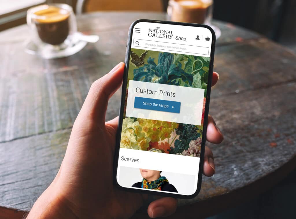 A hand holding a smartphone displaying The National Gallery Shop's website