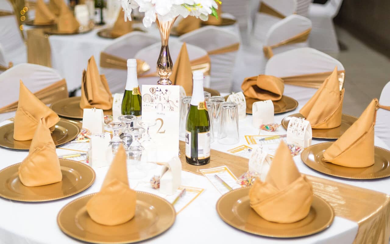 Table covered in white fabric decorated with gold plates, gold napkins, a gold table runner, champagne bottles and flowers in the middle on the table and accessories.