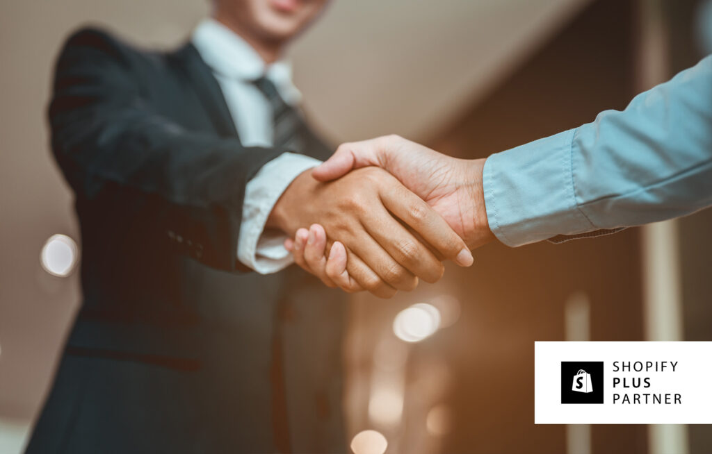 A person in a suit shaking hands with another person, their arm is in shot and they are wearing a blue shirt.